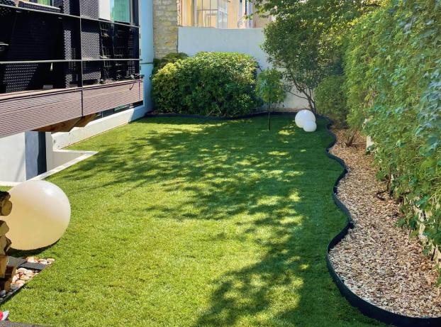 Jardin vu depuis une terrasse accolée à une maison moderne. À gauche, on aperçoit une partie de la maison : un mur blanc avec de grandes fenêtres et un balcon noir en métal.