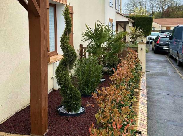 La photo montre l’aménagement paysager d’un petit jardin en façade de maison, le long d’une rue résidentielle. À gauche, on voit la maison avec une façade claire et des éléments en bois foncé autour des fenêtres et des poteaux de soutien du porche.