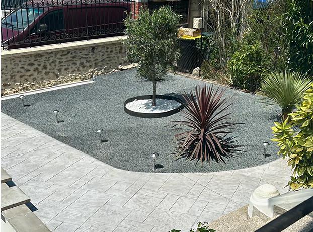 La photo montre un jardin d’entrée très minéral et moderne, vu en légère hauteur, comme depuis un perron ou un escalier. Au premier plan, on distingue une grande surface en dalles grises claires, posées de façon régulière, formant une terrasse ou une allée.