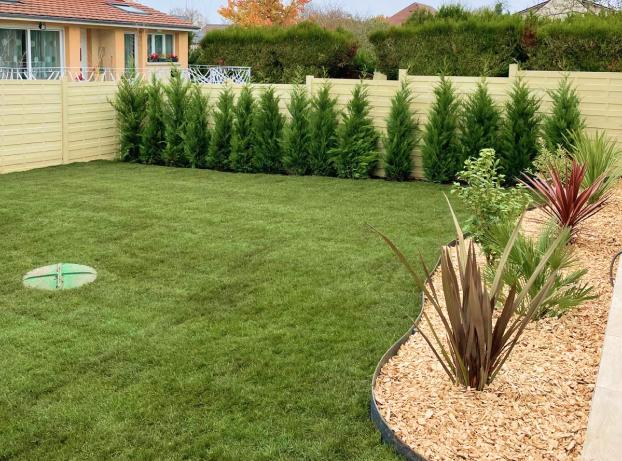La photo montre un jardin arrière, propre et récemment aménagé, vu depuis une terrasse située à droite. Au premier plan à droite, un massif de plantation en forme arrondie est recouvert de copeaux de bois clairs.