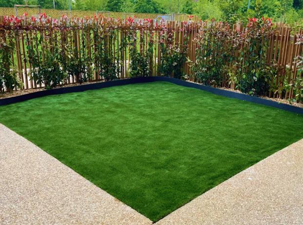La photo montre un petit jardin moderne vu depuis une terrasse en revêtement clair et granuleux. Au centre, une pelouse verte, dense et parfaitement régulière forme un carré légèrement arrondi dans les angles.